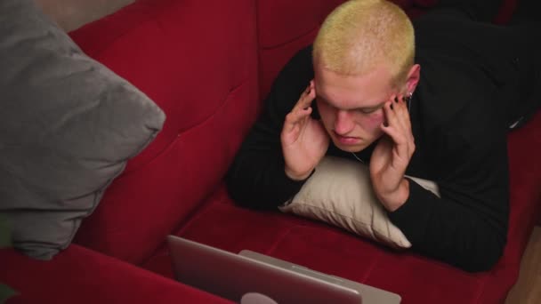 De cerca. Retrato de un guapo joven homosexual cansado usando un portátil, viendo contenido, teniendo dolor de cabeza en el sofá en casa. Hombre con manicura frotando las sienes, teniendo estrés. — Vídeos de Stock