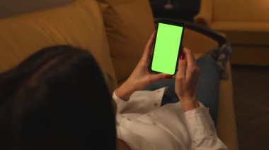 Arkadan bak. Ofisteki kanepede uzanan telefon yeşili model kromakey kullanan bir kadın. Akıllı telefon kullanan, interneti gezen, içeriği izleyen bir kız. Kadının telefonda görüntülü konuşması var..