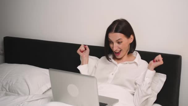 Mujer emocionada tiene buenas noticias usando el ordenador portátil en el dormitorio. Mujer sonriente trabajando en el portátil. Ganador alegre celebrando la victoria en la lotería en línea. — Vídeo de stock