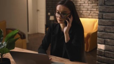 Kendine güveni tam, gözlüklü beyaz bir kadın evde ya da ofiste bilgisayarla telefonda konuşuyor. Masada oturan odaklanmış iş kadını evde dizüstü bilgisayarla çalışıyor..