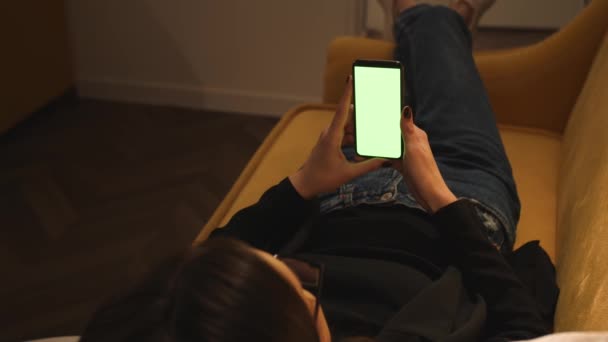 Vedere înapoi. Femeie folosind telefon verde mock-up ecran cromakey culcat pe canapea canapea la birou acasă. Fata care utilizează smartphone-ul, Navigarea pe Internet, Vizionarea conținutului. Femeia are un apel video chat la telefon . — Videoclip de stoc