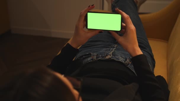 In Großaufnahme halten Frauenhände ein Smartphone mit grünem Chromakey-Bildschirm, das Inhalte anschaut, und wischen auf dem heimischen Sofa herum. Gadgets Menschen Konzept.