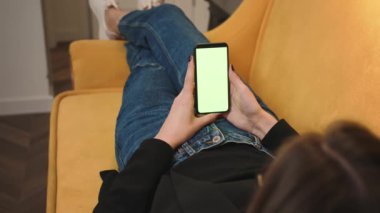 Arkadan bak. Ofisteki kanepede uzanan telefon yeşili model kromakey kullanan bir kadın. Akıllı telefon kullanan, interneti gezen, içeriği izleyen bir kız. Kadının telefonda görüntülü konuşması var..