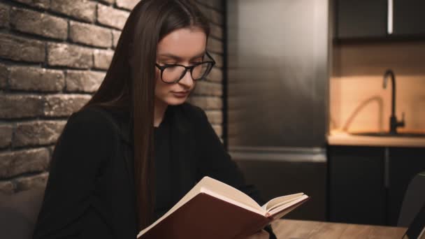 Una joven guapa con gafas leyendo un libro en casa. Estudiante con gafas lee un libro, sentada en casa. — Vídeos de Stock