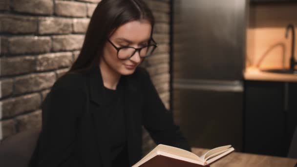 Una joven guapa con gafas leyendo un libro en casa. Mujer sonriente estudiante en gafas lee un libro, sentado en casa. — Vídeo de stock