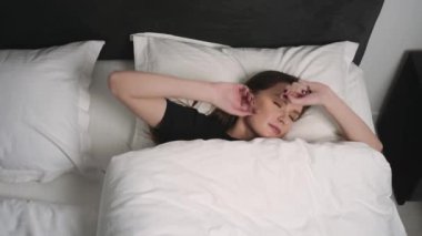  Rahatlamış gülümseyen kadın portresi yatakta beyaz bir yastığın üzerinde uyanır. Evdeki yatak odasında dinlenen beyaz bir kadının portresi. Boş zaman ve rahatlama..