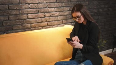 Gözlüklü, telefon kullanan, müzik dinleyen, evdeki kanepede dans eden güzel beyaz bir kadın. İçeride. Evde kal..