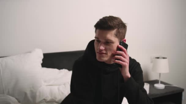 Jóképű kaukázusi férfi beszél otthon telefonon. Egy férfi ül az ágyon és telefonál.. — Stock videók