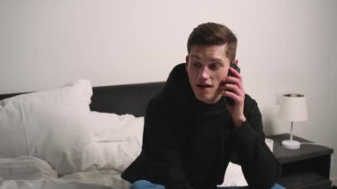 Gülümseyen yakışıklı beyaz adam evde telefonla konuşuyor. Adam yatakta oturuyor ve telefonda konuşuyor..