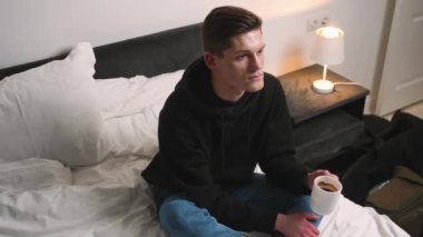 Yakışıklı, gülümseyen adam yatakta oturup kahve içiyor. Yatakta kahve içen, içeriği izleyen, TV 'de film izleyen bir adam..