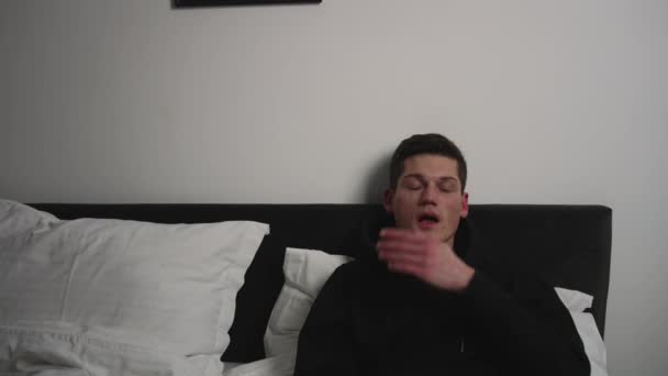 Tired young caucasian man sitting on the bed late at night, yawning and falling asleep. — Stock Video