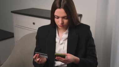 Elinde kredi kartı ve akıllı telefon olan gülümseyen genç bir kadın evdeki ya da ofisteki koltukta oturuyor. İnternetten alışveriş yapan mutlu bayan alışverişçi kolay taşınabilir ödemeler yapıyor. E-bankacılık uygulaması