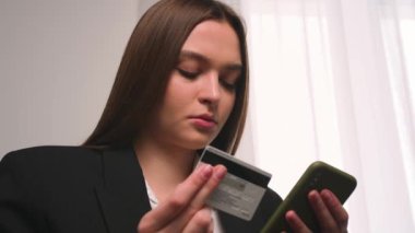 - Yakın çekim. Kredi kartı ve akıllı telefon taşıyan genç bir kadının portresi evdeki ya da ofisteki koltukta oturuyor. İnternetten alışveriş yapan kadın kolay taşınabilir ödemeler yapıyor. E-bankacılık uygulaması