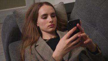 Rahat bir kız elinde akıllı telefonuyla gezici uygulamalar kullanıyor video izliyor, kanepede uzanıyor, tembel genç bir kadın sosyal medyada evdeki kanepede dinleniyor..