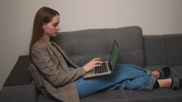 Kaukasische hübsche junge Frau tippt auf Laptop mit grünem Bildschirm, auf dem Sofa zu Hause liegend. Chromakey