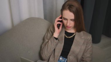 Bilgisayarda çalışırken cep telefonuyla konuşan odaklanmış iş kadınının portresi. Genç bir kadın ev ofisinde dizüstü bilgisayar kullanıyor...
