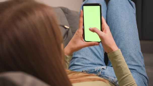 클로즈업. 그린 스크린 스마트 폰 과 소파에 누워 있는 여성. 소녀는 녹색 화면 휴대 전화를 보유하고 있다. 스마트 폰 크로마 키를 사용하는 여성,스위 핑, 스크롤. 인터넷 검색, 비디오 콘텐츠 시청 — 비디오