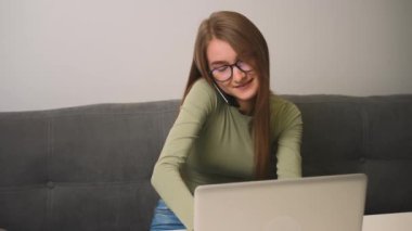 Ofis masasındaki koltukta otururken bilgisayarla konuşan gözlüklü, gülümseyen genç kadın..