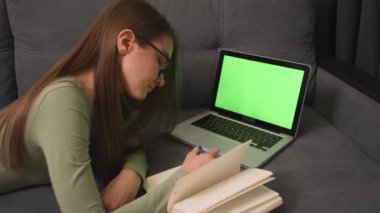 Yeşil ekran krom dizüstü bilgisayar konsepti: online uzaklık eğitimi, video konferansı. Kanepe üzerinde yatan kadın öğrenci evdeki bir deftere bilgi yazıyor..