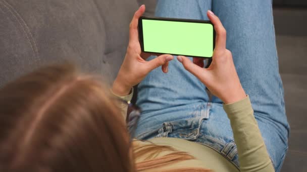 Primer plano. Mujer acostada en un sofá con Smartphone de pantalla verde. Chica sostiene el teléfono móvil de pantalla verde. Mujer usando cromakey Smartphone, Navegación por Internet, Ver contenido de vídeo, Blogs. — Vídeo de stock