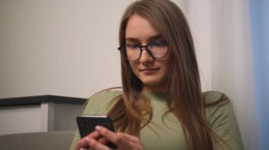 - Yakın çekim. Gözlüklü, gülümseyen çekici genç bir kadının portresi sosyal medyada online alışveriş için telefon kullanıyor koltukta oturuyor, teknolojili mobil mesaj iletişimi yavaş çekim