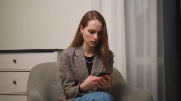 Αναστατωμένη νεαρή γυναίκα κάθεται στον καναπέ, κρατώντας smartphone στα χέρια, έλαβε μήνυμα με άσχημα νέα. Αγχωμένη κυρία σκέφτεται για τα προβλήματα ή τις σχέσεις χωρίζουν μετά την ανάγνωση sms στο τηλέφωνο. — Αρχείο Βίντεο