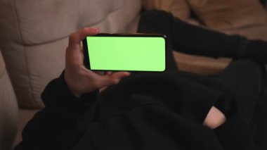 - Yakın çekim. Evdeki Adam Yeşil Model Ekranlı Akıllı Telefon kullanarak Kanepede Uzanıyor, Adam Cep Telefonu Chromakey kullanarak İçeriği İzliyor, İnternet Sosyal Ağ Taraması.