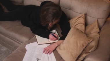 Üst Manzara. Adam evde ders çalışıyor, defter görevleri yazıyor, evdeki kanepede yatıyor..