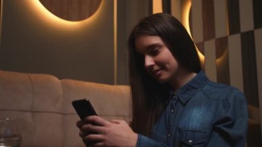 Gülümseyen genç bir kadının portresi, sosyal medyada online alışveriş için telefonu kullanıyor, evdeki koltukta oturuyor, teknolojik mobil mesaj iletişimi yavaş çekim