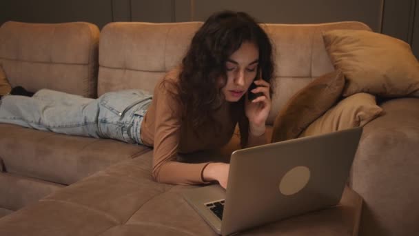 Genç bir kadın kanepede uzanıp dizüstü bilgisayar kullanırken telefonla konuşuyor, internette geziniyor sosyal medyada çalışıyor ya da internette çalışıyor.. — Stok video
