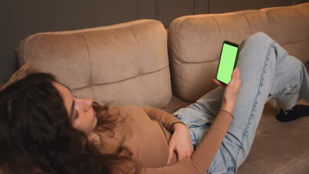 Женщина лежит на диване со смартфоном с зеленым экраном. Девушка держит зеленый экран мобильного телефона. Женщина использует хромаки смартфонов, свинг, прокрутка. просмотр Интернета, просмотр видео контента, блоги. — стоковое видео