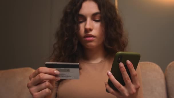 閉鎖だ。自宅やオフィスのソファに座ってクレジットカードやスマートフォンを保持する若い女性の肖像画。オンラインストアで購入する即時簡単なモバイル決済を使用して女性買い物客。電子銀行アプリ — ストック動画