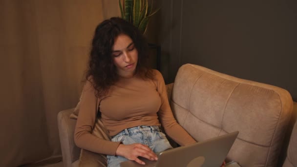 Genç bir kadın kanepede oturup dizüstü bilgisayar kullanıyor, internette geziniyor sosyal medyada çalışıyor ya da evde çalışıyor.. — Stok video