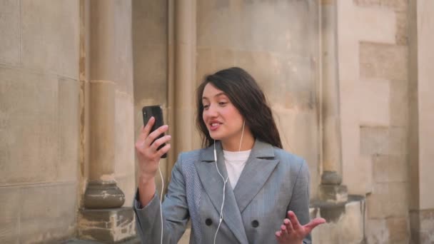 イヤホンを身に着けている若い女性スマイリングトークビデオ通話を作ります,街の背景にリラックスクローズアップ. — ストック動画