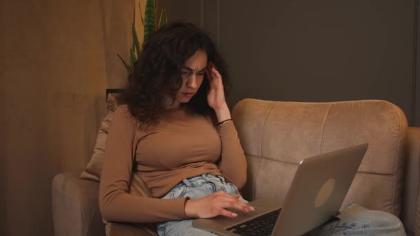 Kanepede oturup dizüstü bilgisayar kullanan, internette gezinen sosyal medyada okuyan ya da evde çalışan genç ve yorgun bir kadın.. — Stok video