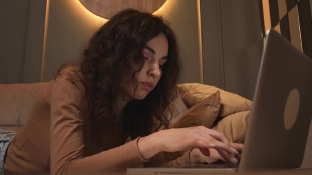 Nahaufnahme. Porträt einer jungen Frau, die mit dem Laptop auf der Couch liegt und auf den Bildschirm schaut, während sie zu Hause im Internet surft, studiert oder online arbeitet. — Stockvideo