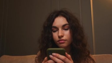 - Yakın çekim. Gülümseyen genç bir kadının portresi, sosyal medyada online alışveriş için telefonu kullanıyor, evdeki koltukta oturuyor, teknolojik mobil mesaj iletişimi yavaş çekim