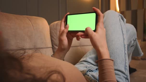 Primer plano. Mujer acostada en un sofá con Smartphone de pantalla verde. Chica sostiene el teléfono móvil de pantalla verde. Mujer Usando Smartphone chromakey, deslizando, desplazándose. navegar por Internet, ver contenido de vídeo — Vídeos de Stock