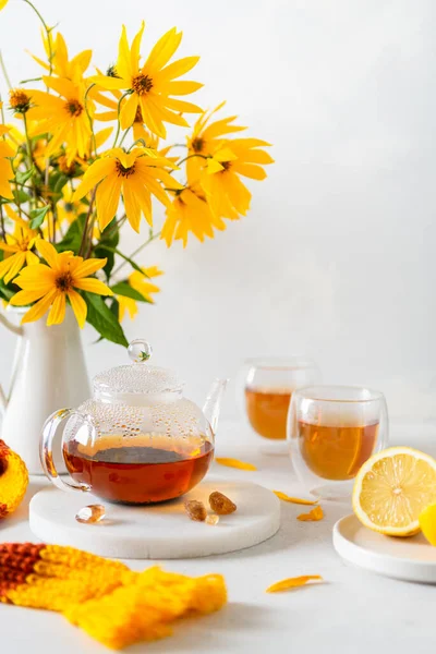 Cozy autumn. Fall composition, yellow flowers bouquet, hot teapot, two cups of tea and a warm scarf on white table background. Tea party. Poster, greeting card. Copy space for text
