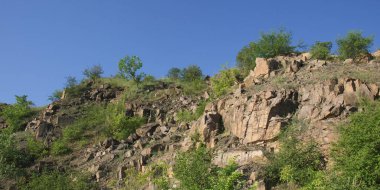 Sıcak bir yaz gününde Ukrayna 'nın Nikolaev bölgesinde terk edilmiş bir granit ocağının kayalık yamacı. Keskin taşlar seyrek bitkilerle kaplanmış. Uçurumların üzerinde bulutsuz mavi gökyüzü.