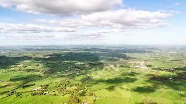 Yükseltilmiş bir perspektif geniş tarım arazilerini gösterir. Yeşil çayırlar ve tarlalar yeryüzü boyunca uzanıyor. Kabarık beyaz bulutlarla dolu bir gökyüzü altında, yerde gölgeler yaratıyor..