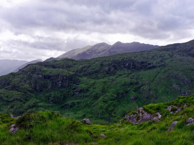 Serene mountain landscape bathed in soft sunlight. Rolling green hills stretch towards majestic peaks under a partly cloudy sky. Ideal for travel, nature, and adventure themes.