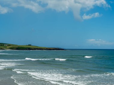 This is a view of the ocean on a sunny day in West Cork. Waves are moving toward the sandy beach. Grass and a house are visible along the coastline in Ireland.
