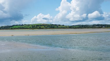 Ufukta süzülen yeşil tepeler ve yumuşacık bulutlarla dolu bir gökyüzünün altında kumlara akan huzurlu bir su manzarası..