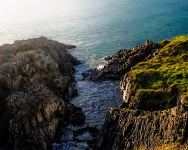 Okyanus dalgaları, sivri ve karanlık uçurumlar arasında dar bir geçitten geçer. Batı Cork, İrlanda 'daki kayalıkların bir kısmını yemyeşil çimenler kaplıyor.