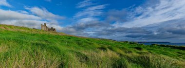 Yeşil çimenli bir yamaç, Batı Cork 'taki okyanusa doğru iniyor. Harabe binalar tepenin tepesinde, beyaz ve gri bulutlarla dolu bir gökyüzünün altında duruyor..