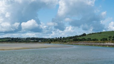 Suyun kıyılarla buluşmasının güzelliği, gökyüzünün altında yeşil tepeler tarafından desteklenen yumuşak, kabarık bulutlar, huzurlu bir manzara..