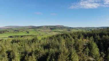 A sunny day. Green fields and dense forest extend to the horizon. Distant wind turbines are visible on the hills.