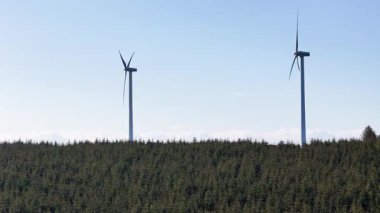 Rüzgâr türbinleri yoğun bir ormanın üzerinde açık mavi gökyüzüne karşı yükselirler, yenilenebilir enerji ve çevresel sürdürülebilirliği sembolize ederler..