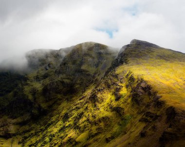 Bu manzara Coomloughra At nalı Dağları 'nın engebeli arazisini gösteriyor. Yeşil çimenler yamaçları kaplıyor ve yürüyüş yolları açıkça görülüyor. Bulutlar dağ zirvelerini kısmen karartıyor.